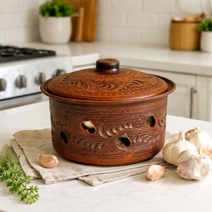 Könnte beinhalten: Ein brauner Knoblauchbehälter aus Keramik mit Deckel, dekorativen Aussparungen und einer strukturierten Oberfläche. Der Behälter ist mit Knoblauchzehen gefüllt und liegt auf einem Leinentuch, mit ganzen Knoblauchknollen in der Nähe. Eine Küche.