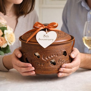 Könnte beinhalten: Ein brauner Keramiktopf mit Deckel und orangefarbenem Band. Der Topf hat dekorative Ausschnitte und ein herzförmiges Etikett mit der Aufschrift "8th Anniversary". Der Topf wird von zwei Personen gehalten.
