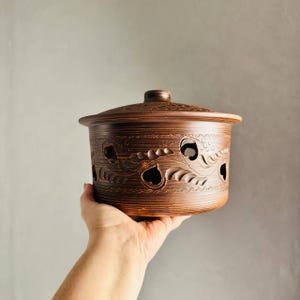 Könnte beinhalten: Ein brauner Keramiktopf mit Deckel, gehalten in einer Hand. Der Topf hat dekorative Ausschnitte und geätzte Blumenmuster. Der Topf hat einen Durchmesser von etwa 15 cm und ist 10 cm hoch.