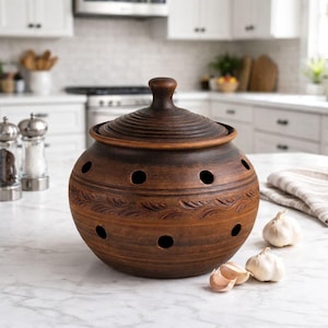 May include: A brown ceramic garlic keeper with a lid, featuring decorative leaf patterns and circular ventilation holes. The keeper is designed to store garlic and keep it fresh. Several garlic cloves are placed near the keeper.