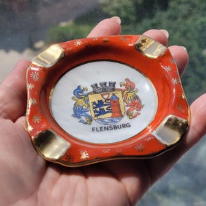 Puede incluir: Un pequeño cenicero de cerámica roja con detalles dorados. El centro presenta un óvalo blanco con un escudo de armas colorido y la palabra "FLENSBURG" debajo. El borde está decorado con estrellas doradas.