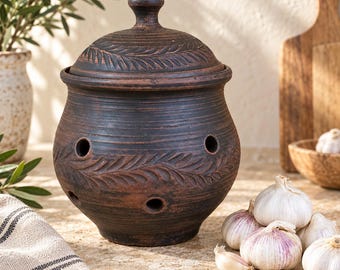 May include: A dark brown, round ceramic garlic keeper with a matching lid. The vessel features a horizontal, carved leaf pattern around the center and multiple circular ventilation holes. The surface has a textured, matte finish with subtle variations.