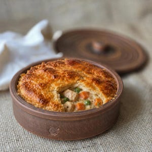 Puede incluir: Un pastel de carne salado con una corteza de hojaldre dorada se encuentra dentro de un plato redondo de cerámica marrón mate. El relleno contiene trozos visibles de carne, zanahorias de color naranja brillante y guisantes verdes en una salsa cremosa. Una tapa a juego descansa cerca.