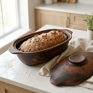 May include: An oval-shaped dark brown clay baking dish with two side handles, containing a golden-brown loaf of crusty bread. The matching lid, featuring an embossed floral pattern, rests beside the dish on a light-colored fabric cloth.
