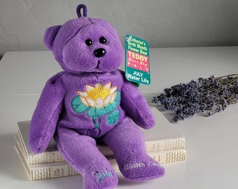 May include: A purple plush teddy bear with a loop on its head, featuring an embroidered water lily on its chest. The bear has the word July stitched on one foot and Water Lily on the other. A green tag reads Collector's Birth Month Flower Bear.