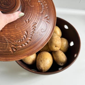 以下が含まれることがあります： 茶色の陶器のボウルと蓋で、ジャガイモが詰まっています。蓋には浮き彫りの花の模様があります。ボウルには装飾的な切り欠きがあります。ジャガイモは薄茶色で楕円形です。