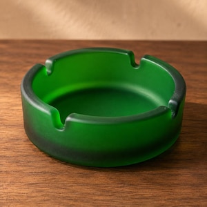 May include: A round, emerald green glass ashtray with a frosted, matte finish. The circular vessel features four evenly spaced notches along the thick, raised rim, designed to hold items securely in place while maintaining a smooth, uniform profile.