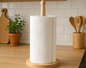 May include: A light-colored wooden paper towel holder with a cylindrical base and a central vertical rod topped with a rounded knob. A roll of white paper towels with a repeating embossed geometric star pattern is mounted on the wooden stand.