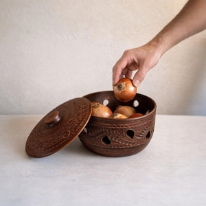 Peut inclure: Un bol de rangement rond en argile brune avec un couvercle assorti, présentant des motifs décoratifs sculptés et plusieurs trous de ventilation en forme de goutte d'eau sur les côtés. Le bol contient plusieurs oignons jaunes entiers.