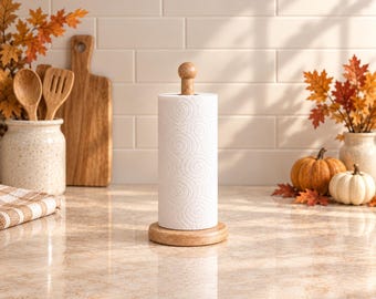 Puede incluir: Un soporte para papel de cocina con una base circular de madera marrón claro y una varilla vertical a juego rematada con una pequeña esfera. Un rollo de papel de cocina blanco con un patrón de espirales en relieve está en el soporte.