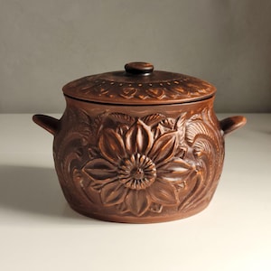 以下が含まれることがあります： ひまわりのデザインが施された、蓋付きの茶色の陶器の鍋。鍋には2つの取っ手と蓋に小さなつまみがあります。ひまわりは中心的なデザイン要素で、詳細な彫刻が施された花びらがあります。鍋は調理または提供に使用されます。