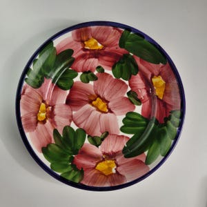 以下が含まれることがあります： 白いベースに手描きの花柄が施された丸いセラミックプレート。花はピンク色で、中心は黄色、葉は緑色です。プレートには濃い青色の縁があります。
