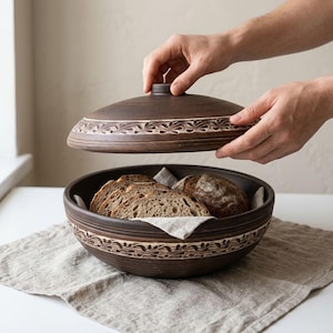 Puede incluir: Una panera de cerámica marrón oscuro con tapa a juego, con un diseño tallado decorativo. La cesta contiene pan en rodajas y una hogaza entera, sobre un paño de lino. Se levanta la tapa, revelando el contenido.