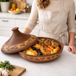 May include: A brown ceramic tagine with a decorative lid, filled with cooked food including chicken, carrots, and couscous. The tagine is open, revealing the meal inside. The dish is on a white countertop.