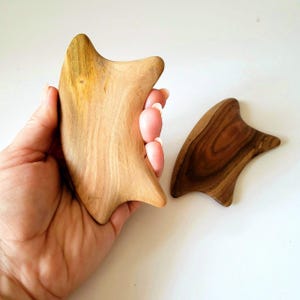 May include: Two smooth, irregularly shaped wooden massage tools with curved edges. One tool is a light tan colour with visible wood grain, while the other is a darker brown with prominent, swirling grain patterns. Both have a contoured design.