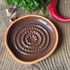 May include: A round, brown ceramic grater plate with a raised, flower-shaped texture in the center for grating garlic or ginger. The plate features a smooth, glazed interior surface and an unglazed, light-colored terracotta rim edge.