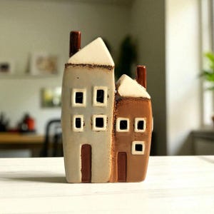 Könnte beinhalten: Ein Set dekorativer Keramik-Häuser. Eines ist hellgrau, das andere braun. Beide haben weiße Dächer, dunkelbraune Schornsteine und quadratische Fenster. Die Häuser sind als dekorative Akzente konzipiert.