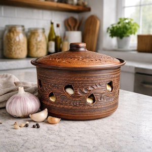 Könnte beinhalten: Ein brauner Keramik-Knoblauchbehälter mit Deckel, mit dekorativen blattartigen Aussparungen und einer strukturierten Oberfläche. Der Behälter ist mit Knoblauchzehen gefüllt und steht auf einer Arbeitsplatte.