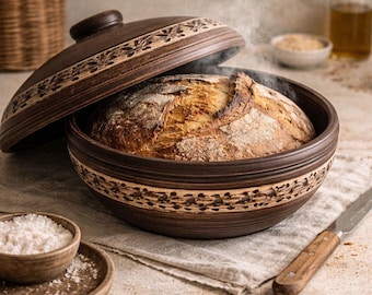 Puede incluir: Una olla de barro redonda de color marrón oscuro con tapa a juego, que presenta un intrincado patrón floral tallado alrededor del borde. Dentro de la olla hay una hogaza de pan dorada recién horneada con vapor visible saliendo.