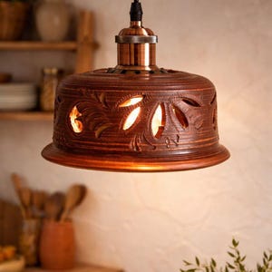 May include: A brown ceramic pendant light fixture with a wide, dome-shaped shade featuring intricate cut-out leaf patterns that allow warm light to shine through. The lamp is suspended by a black cord from a metallic copper-colored hardware fitting.