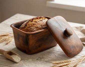 May include: A rectangular brown clay bread baker with a matching lid, containing a golden-brown loaf of bread. A small wooden scoop filled with white flour and several stalks of dried wheat are positioned near the container.