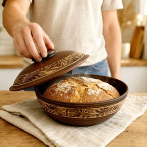 May include: A round, dark brown clay baking bowl with a matching lid, both featuring intricate, carved floral patterns. Inside the bowl sits a freshly baked, golden-brown loaf of crusty bread with a dusted flour top and visible steam rising.