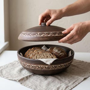May include: A dark brown ceramic bread bowl with a matching lid, featuring a carved leaf pattern around the rim. The bowl contains several slices of crusty sourdough bread resting on a light beige linen cloth, with the lid being lifted.