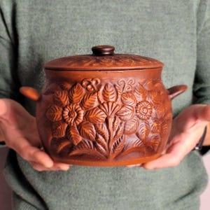 May include: A brown ceramic pot with a lid, decorated with a floral relief design. The pot has two handles and a knob on the lid. The floral pattern includes detailed leaves and flowers. The pot is held by a person wearing a green jumper.