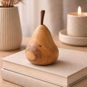 Pode incluir: Um objeto decorativo em forma de pera esculpido em madeira castanho-clara com um acabamento liso e polido e um caule de madeira castanho-escura. O objeto apresenta veios de madeira natural visíveis e uma base arredondada sobre dois livros.