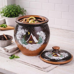 May include: A round, dark brown ceramic crock with a matching lid, featuring a hand-painted winter scene of a cabin in the snow with a yellow crescent moon. The exterior has a speckled finish, and the interior is filled with various food items.