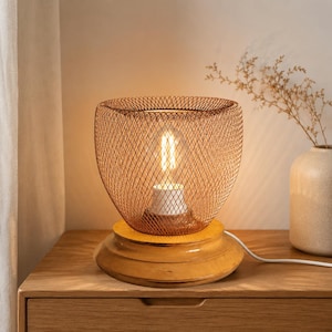 May include: A copper wire mesh lamp with a warm bulb. The lamp sits on a wooden base with a tiered design. The lamp is plugged in. A vase with dried flowers is in the background. The lamp is on a wooden bedside table.