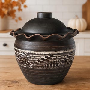 May include: A dark brown ceramic jar with a matching lid and a ruffled rim. The body of the jar features a horizontal band with a white-accented leaf pattern carved into the clay. The surface has a textured, matte finish with visible horizontal lines.