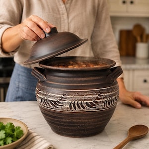 May include: A dark brown, round clay pot with a matching lid and two side handles. The pot features a wide, textured band around the center with a white, leaf-like pattern. Steam rises from the pot, which is filled with a cooked bean dish.