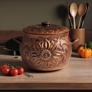 Può includere: Una casseruola in ceramica marrone con coperchio, con un design a girasole. Il piatto ha due manici ed è circondato da cucchiai di legno, pomodori e una piccola zucca. Il tema generale è l'utensileria da cucina.
