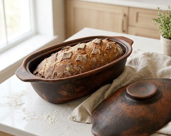 May include: A dark brown oval ceramic baking dish with integrated side handles, featuring an embossed floral pattern on the exterior. A golden-brown loaf of crusty bread sits inside the dish, while the matching lid rests nearby on a cloth.