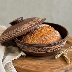 Puede incluir: Un recipiente de barro redondo de color marrón oscuro con tapa a juego, ambos con intrincados patrones florales tallados. Dentro del cuenco hay una hogaza de pan de masa madre recién horneada, dorada y con una corteza espolvoreada con harina.