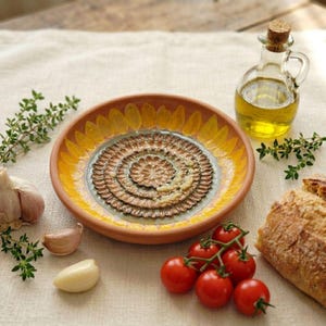 Peut inclure: Une assiette à trempette ronde en céramique avec un bord jaune inspiré d'un tournesol et un centre texturé brun à motif floral. L'assiette est accompagnée d'une burette en verre remplie d'huile d'olive, de brins de thym, d'ail et de pain.
