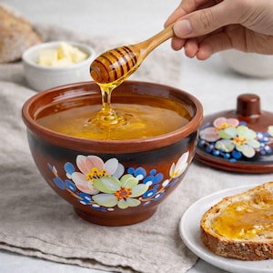 Könnte beinhalten: Eine braune Keramikschale gefüllt mit Honig, mit einem hölzernen Honiglöffel. Die Schale hat Blumenmuster in Weiß, Rosa und Blau. Eine Scheibe Brot mit Honig liegt auf einem Teller. Die Schale hat einen Durchmesser von ca. 15 cm.