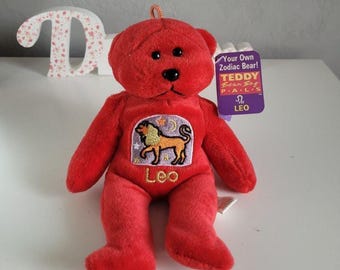 May include: A bright red plush teddy bear featuring a small loop on top of its head. The bear has a patch on its chest depicting a lion with the text Leo below it. A purple tag attached to the ear reads Your Own Zodiac Bear! TEDDY BEAN BAG PALS.