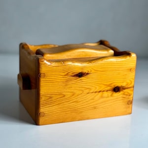 Puede incluir: Una pequeña caja de madera hecha a mano con tapa. La caja está hecha de madera de color claro con vetas y nudos visibles. Tiene forma rectangular con bordes redondeados y un pequeño asa lateral. La tapa tiene un diseño curvo.