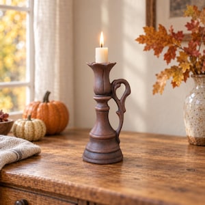 Puede incluir: Un candelabro marrón texturizado con base acampanada, tallo central y un asa curva en el lateral. El soporte presenta una copa superior en forma de flor que sostiene una vela de pilar de color blanco roto encendida con llama amarilla.