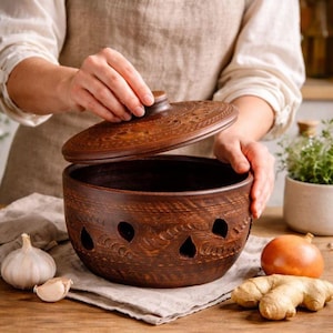 Könnte beinhalten: Ein handgefertigter, brauner Keramik-Knoblauchtopf mit passendem Deckel. Der Topf hat dekorative Aussparungen und ist von frischem Knoblauch, einer Zwiebel und Ingwer auf einer Holzoberfläche umgeben.