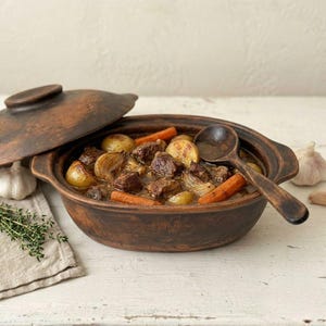 Può includere: Una pirofila in argilla marrone con coperchio abbinato, riempita con uno stufato di pezzi di manzo, carote arrosto e cipolle. Un cucchiaio di legno riposa all'interno del piatto, che presenta un motivo floreale in rilievo sull'esterno e sui manici.