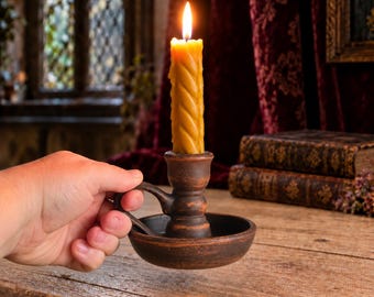 May include: A dark brown, circular clay candle holder with a small handle, featuring a single, tall, yellow, twisted-pattern beeswax candle with a lit flame, held in a hand against a blurred background of books and fabric.