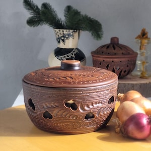 May include: A brown ceramic bowl with a lid, featuring heart-shaped cutouts and decorative carvings. The bowl is part of a kitchenware set, alongside a smaller lidded bowl and a decorative vase. Onions are visible in the foreground.