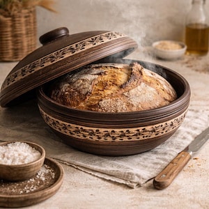 May include: A round, dark brown clay baking dish with a matching lid, featuring an intricate carved floral pattern around the rim. Inside the dish sits a freshly baked, golden-brown loaf of sourdough bread with visible steam rising from the crust.