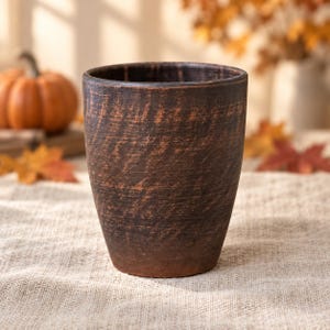May include: A cylindrical cup made of dark brown clay with a visible, textured, and slightly uneven surface. The vessel features a deep, earthy tone with subtle variations in color throughout the exterior, showcasing a raw and unglazed finish.