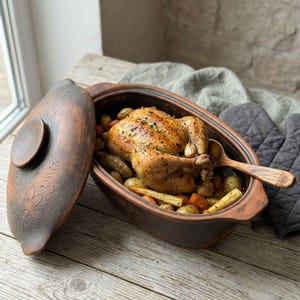 May include: An oval-shaped brown clay roasting pot with a matching lid featuring etched floral patterns. Inside the pot is a roasted whole chicken surrounded by cooked potatoes, carrots, and parsnips, with a wooden spoon resting inside.