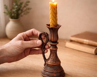 May include: A dark brown ceramic candlestick holder with a handle and embossed floral details, holding a lit yellow beeswax candle with a twisted, braided texture. The candlestick features a flared top and a wide, stable base for support.