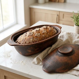 Op de afbeelding: Een bruine keramische broodoven met een vers gebakken brood erin. De ovale bakker heeft een deksel met een bloemenmotief. Het brood heeft een textuurkorst. De bakker staat op een wit aanrecht.
